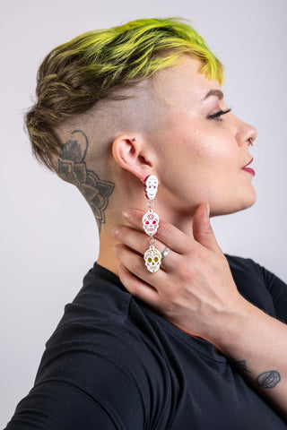 A woman with bright yellow hair and skull tattoo wears the The Bad Button calavera earrings